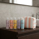 photo professionnelle lifestyle, salle de bain design moderne, ensemble de 5 produits capillaires pour cheveux frisés et bouclés, présentés sur un meuble a côté d'une trousse de voyage en twill blanc rayé rose pêche. 5 flacons de taille small 100ml.